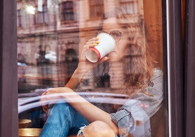 Young redhead curly girl wearing casual clothes and glasses drink a takeaway coffee while sitting on a window sill.