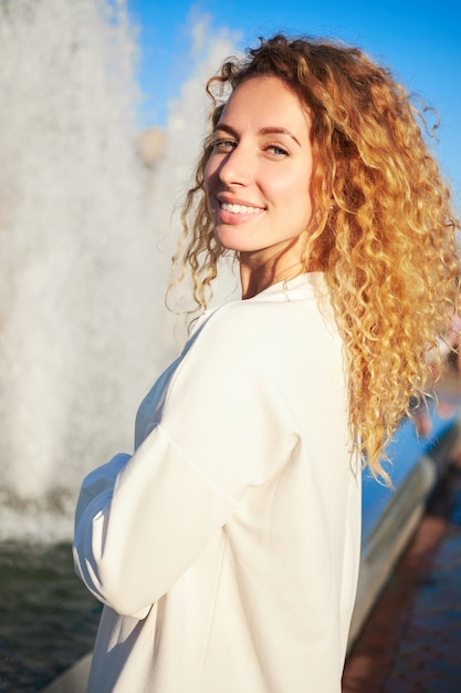 Young redhead curly white smiling girl, weared white dress, stands near fountain