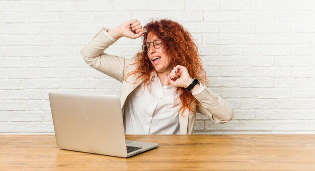 Young redhead curly woman working with her laptop celebrating a special day, jumps and raise arms with energy.