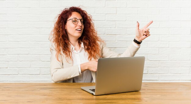 彼女のラップトップで働く若い赤毛の巻き毛の女性は、人差し指で離れて指している興奮しています。