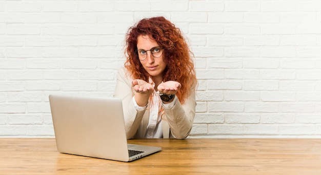 Young redhead curly woman working with her laptop holding something with palms