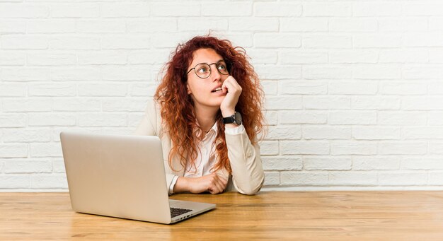 Young redhead curly woman working with her laptop relaxed thinking about something looking at a copy space.