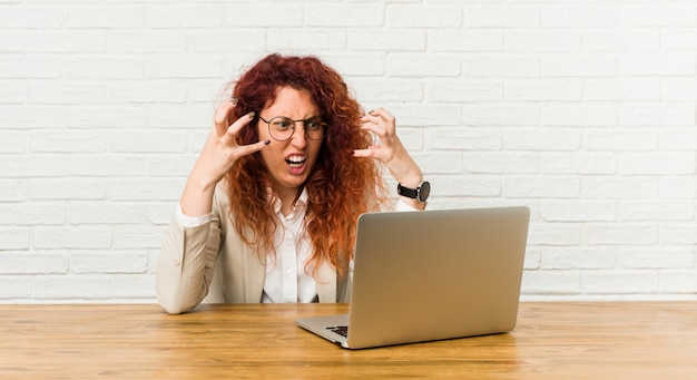 Donna riccia della giovane testarossa che lavora con il suo computer portatile che grida con rabbia