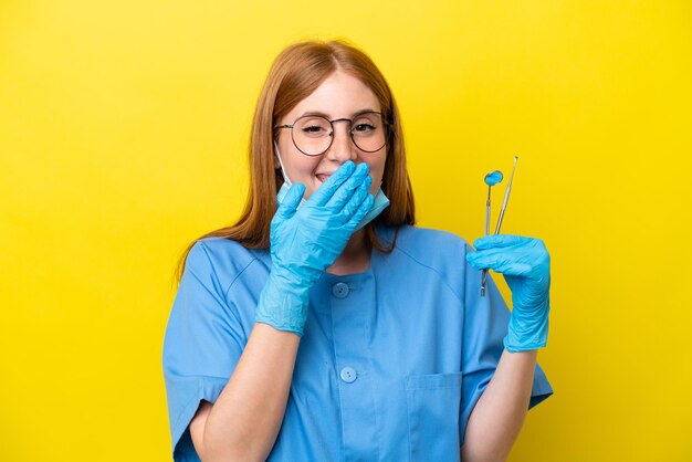 Young redhead Dentist woman isolated on yellow background happy and smiling covering mouth with hand