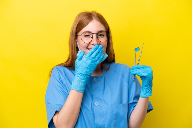 黄色の背景に分離された若い赤毛の歯科医の女性幸せと笑顔の手で口を覆う