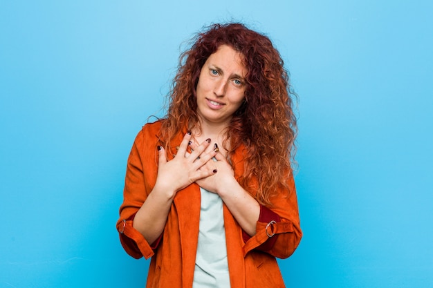 Young redhead elegant woman has friendly expression, pressing palm to chest. Love concept.