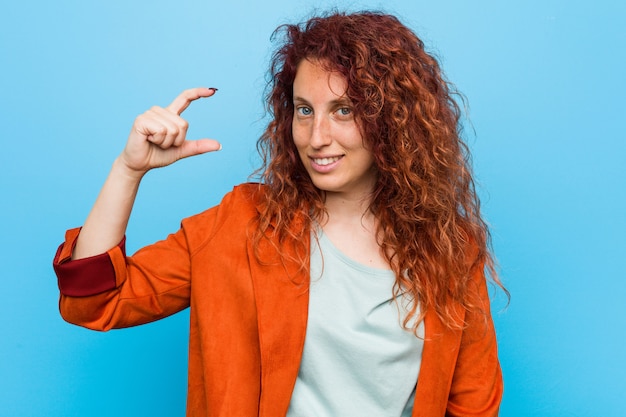 Photo young redhead elegant woman holding something little with forefingers, smiling and confident.