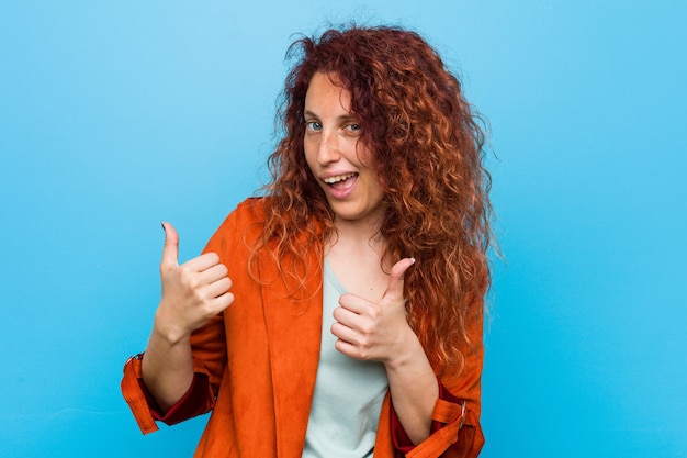 若い赤毛のエレガントな女性は、笑顔と自信を持って、両方の親指を上げます。