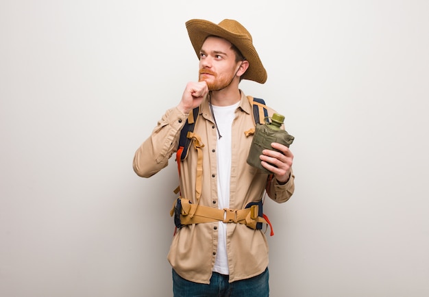 Young redhead explorer man doubting and confused. He is holding a canteen.