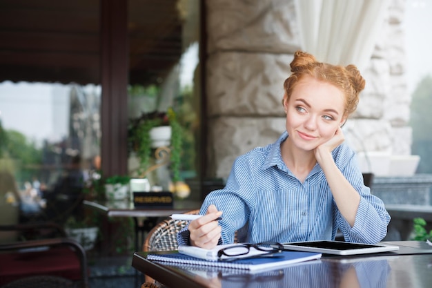 Young redhead female freelancer in glasses making notes in a notebook sitting at summer terrace cafe. Freelancer working businesswoman lifestyle concept.