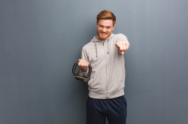 Young redhead fitness man cheerful and smiling pointing to front. Holding a dumbbell.