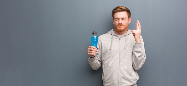 Young redhead fitness man crossing fingers for having luck. He is holding an energy drink.