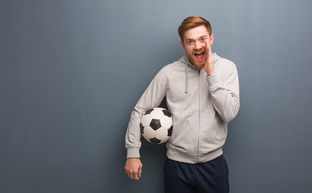 Young redhead fitness man shouting something happy to the front. He is holding a soccer ball.