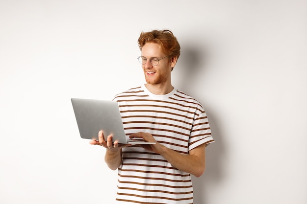 Молодой рыжий фрилансер работает на ноутбуке, печатает на клавиатуре компьютера и улыбается, стоя на белом фоне.