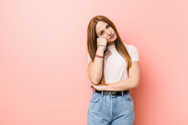 Young redhead ginger woman against a pink wall who feels sad and pensive, looking at copy space.