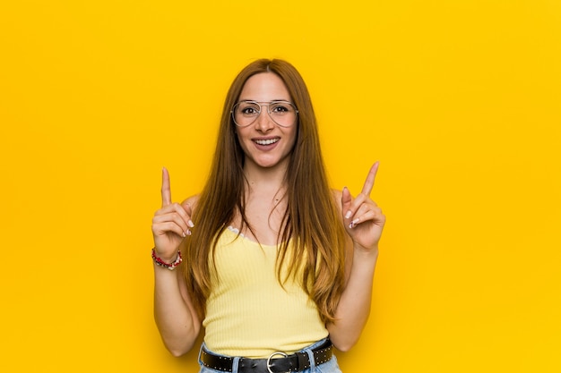 Young redhead ginger woman with freckless indicates with both fore fingers up showing a blank space.