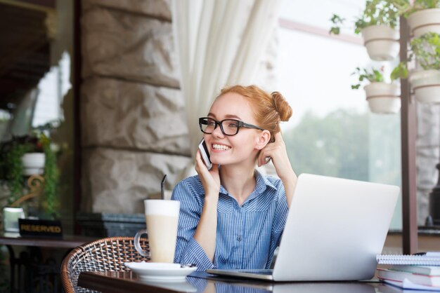 Young redhead girl in glasses talking on phone with laptop, notebooks sitting at summer terrace cafe. Freelancer working businesswoman lifestyle concept.