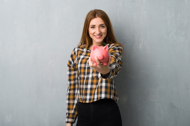 Young redhead girl over grunge wall holding a piggybank