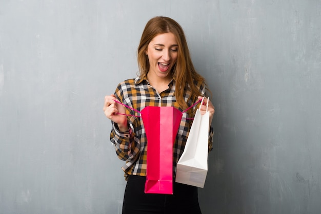 Young redhead girl over grunge wall surprised while holding a lot of shopping bags