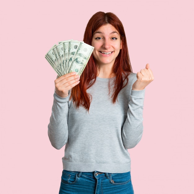 Young redhead girl happy because has won a lot of money on isolated pink background