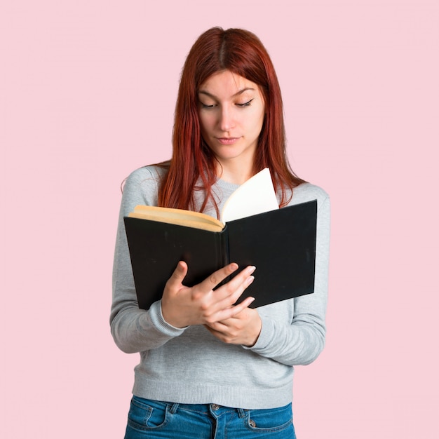 Young redhead girl holding a book and enjoying reading on isolated pink background