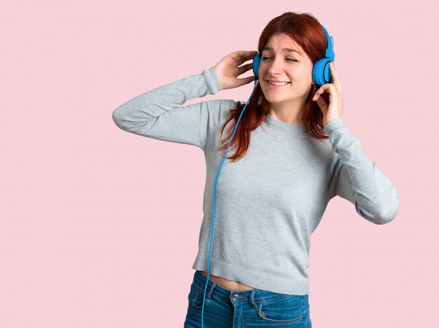 Young redhead girl listening to music with headphones on isolated pink background