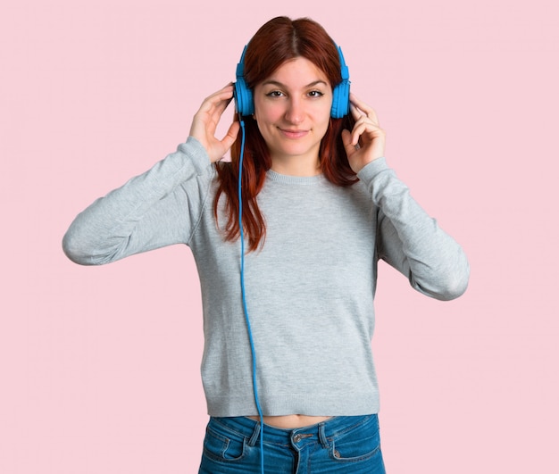 Foto giovane ragazza della testarossa che ascolta la musica con le cuffie su fondo rosa isolato