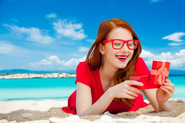 Young redhead girl in red dress with gift box have a rest on summertime sea beach