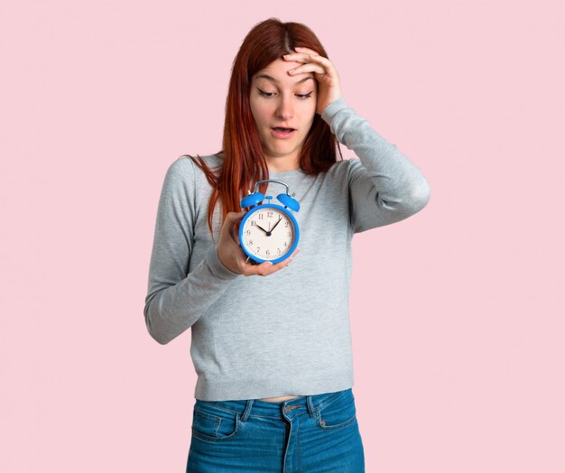 Young redhead girl restless because it has become late and holding vintage alarm clock on isolated p