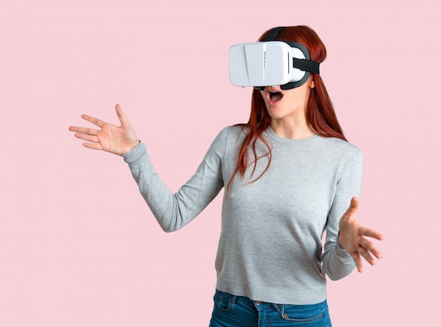 Young redhead girl using VR glasses. Virtual reality experience on isolated pink background