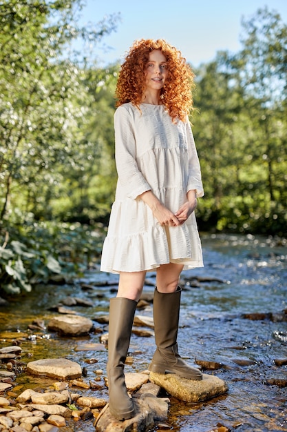 Young redhead lady with curly hairstyle posing at camera, wearing white dress