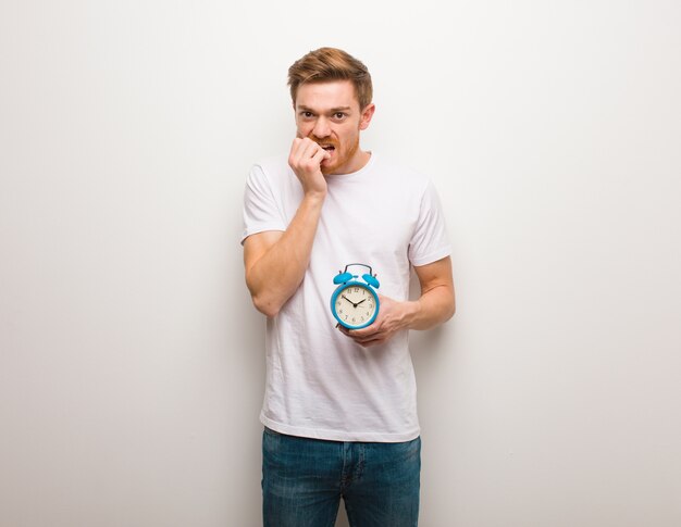 Young redhead man biting nails, nervous and very anxious. He is holding an alarm clock.