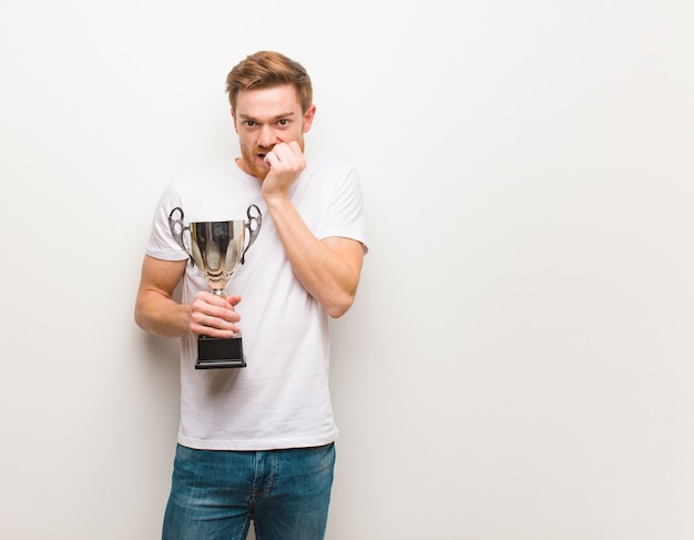 Young redhead man biting nails, nervous and very anxious. Holding a trophy.