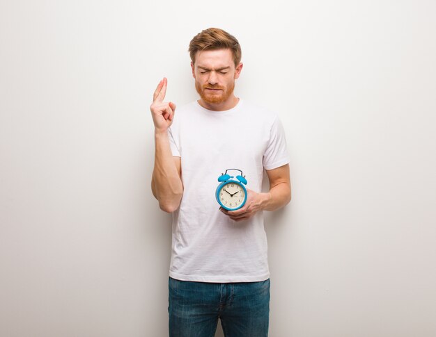 Young redhead man crossing fingers for having luck. He is holding an alarm clock.