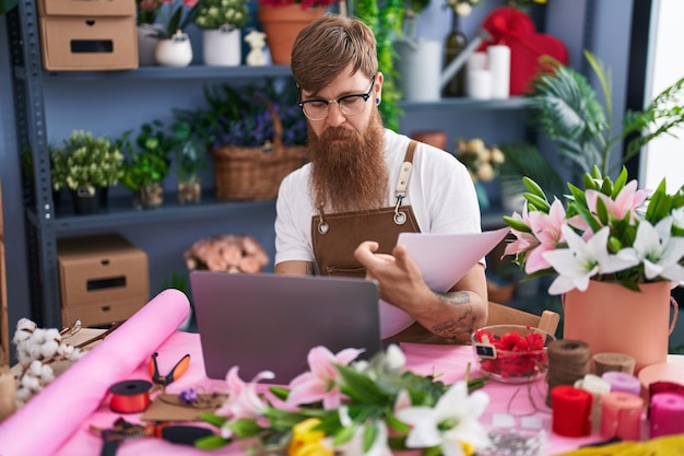 Photo young redhead man florist using laptop reading document at flower shop