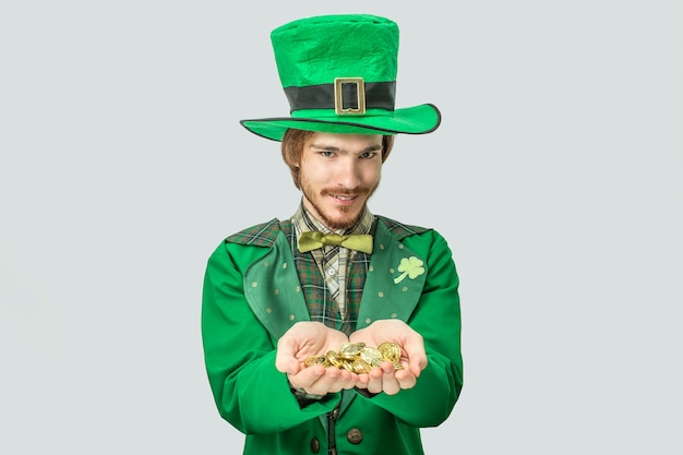 Young redhead man holding lots of golden coins in hands and show them to camera. He look straight forward. Guy wear saint Patrick's suit. Isolated on grey .