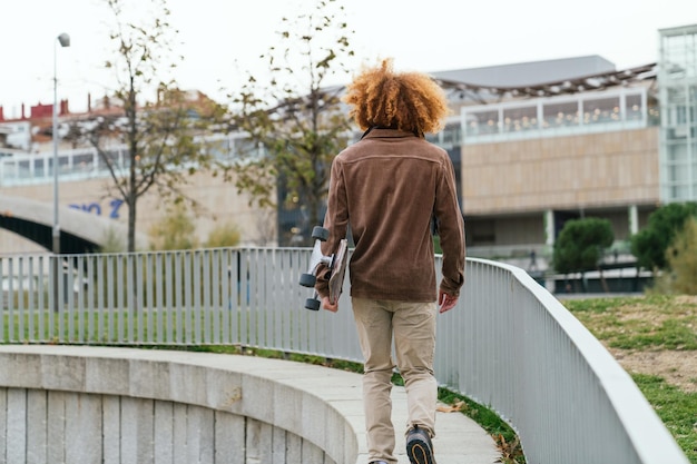 スケートボードを握っている若い赤の男性が都市道を歩いています