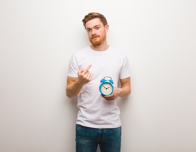 Young redhead man inviting to come. He is holding an alarm clock.