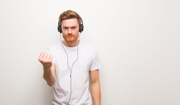 Young redhead man showing fist to front, angry expression. Listening to music with headphones.