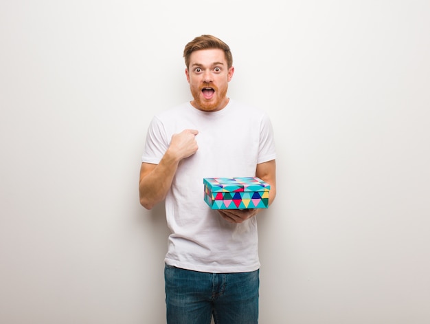 Young redhead man surprised, feels successful and prosperous. Holding a gift box.