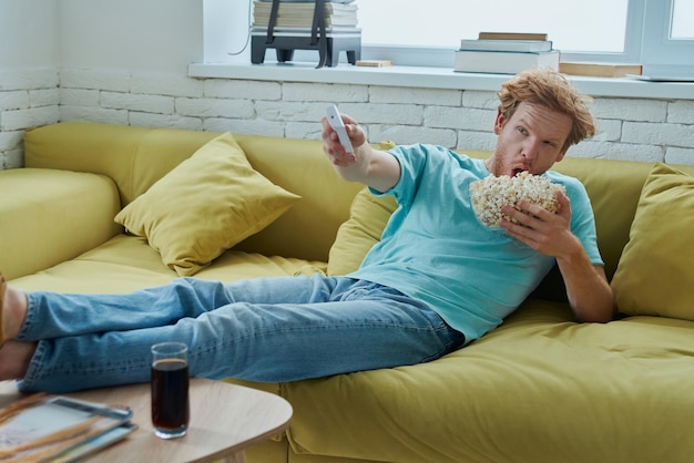 自宅のソファに座ってテレビを見たり、おやつを楽しんでいる若い赤毛の男