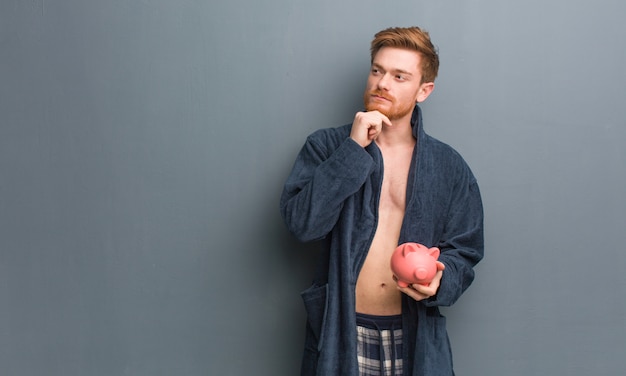 Young redhead man wearing pajama doubting and confused. He is holding a piggy bank.