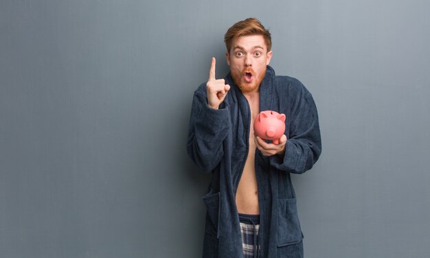 Young redhead man wearing pajama having a great idea,  of creativity. He is holding a piggy bank.