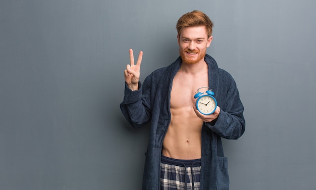 Young redhead man wearing pajama showing number two. He is holding an alarm clock.