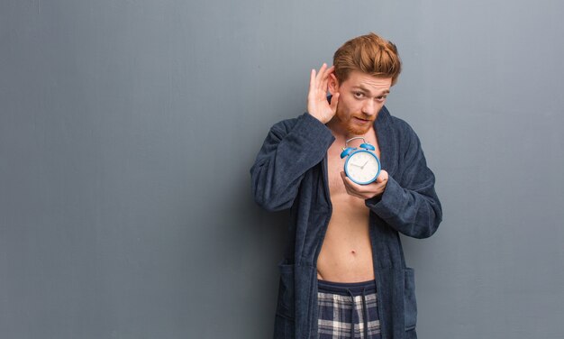 Young redhead man wearing pajama try to listening a gossip. He is holding an alarm clock.