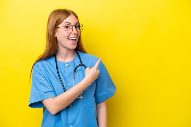 Молодая рыжая медсестра, изолированная на желтом фоне, удивлена и указывая в сторону