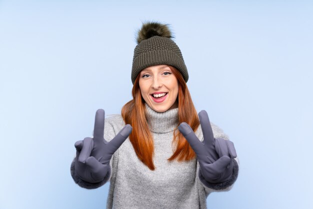 Young redhead Russian woman with winter hat over isolated blue  smiling and showing victory sign