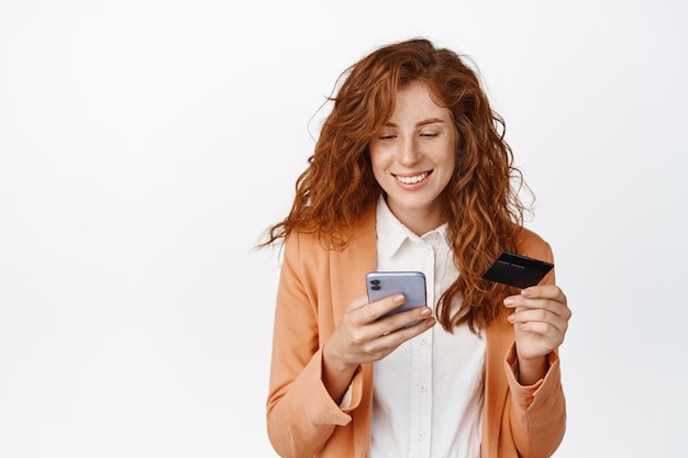 Young redhead saleswoman using mobile phone and credit card Smiling businesswoman paying online with smartphone app standing over white background