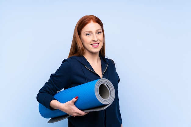 Young redhead sport woman over isolated blue background with a mat and smiling