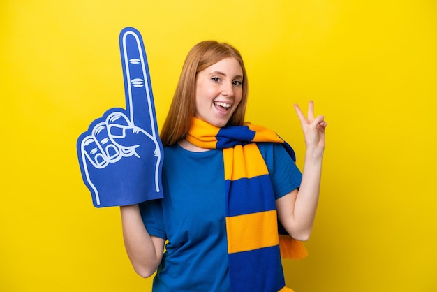 Young redhead sports fan woman isolated on yellow background smiling and showing victory sign